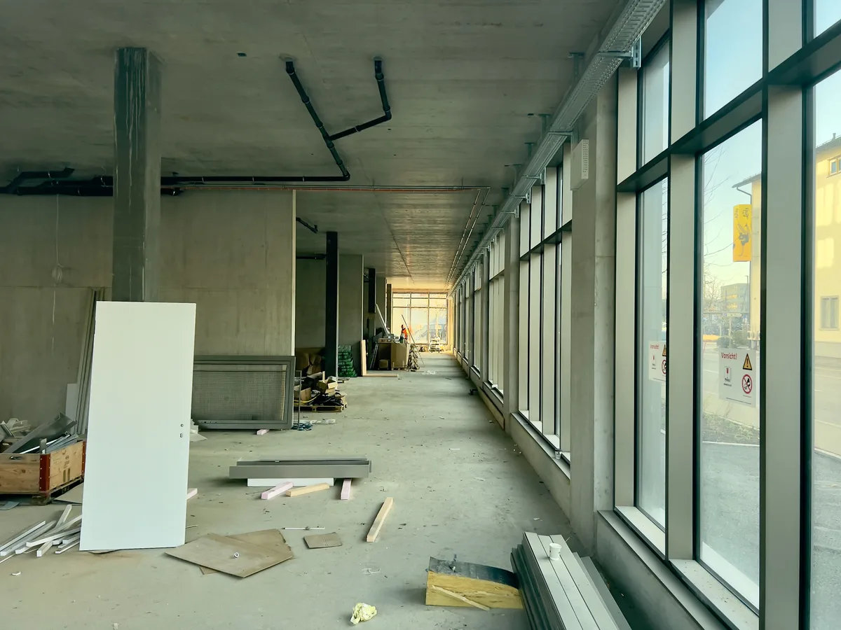 Man sieht durch ein Fenster in einen langen Gang in eines Rohbaus. Verstreut liegt Baumaterial auf dem Boden.